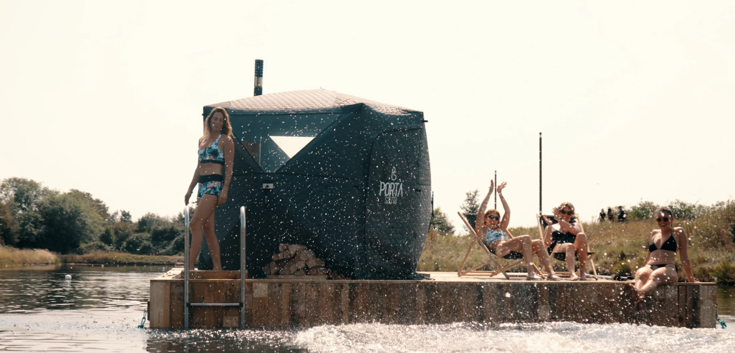 A wood-fired mobile sauna on a floating pontoon, with swimmers relaxing on deck chairs